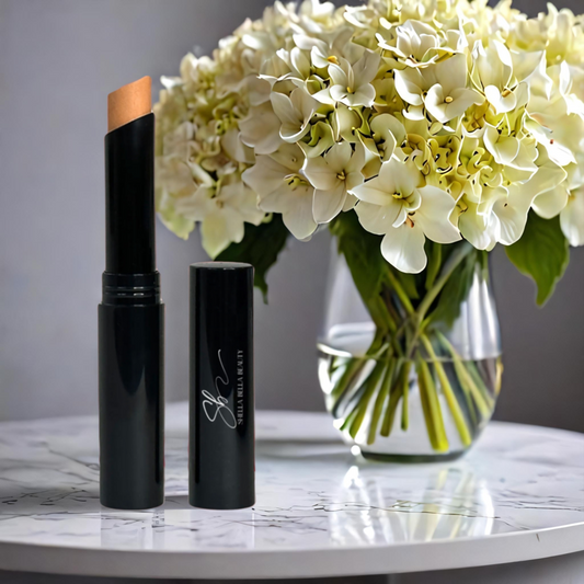 A RadiantBlend™  Butter Creme Concealer Stick, with the cap off, displayed next to its container, against a background featuring a vase of white flowers on a marble surface.