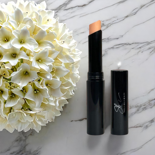 A RadiantBlend™ Almond Creme Concealer Stick next to its cap, displayed on a marble surface with a cluster of white flowers to the side.